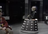 Full-body side-view of Davros as the sceptical Doctor looks on.