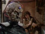 Davros sits in stony silence as the Doctor works on an explosive.